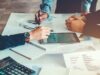 Schreibtisch, auf dem Dokumente liegen und drei Personen arbeiten